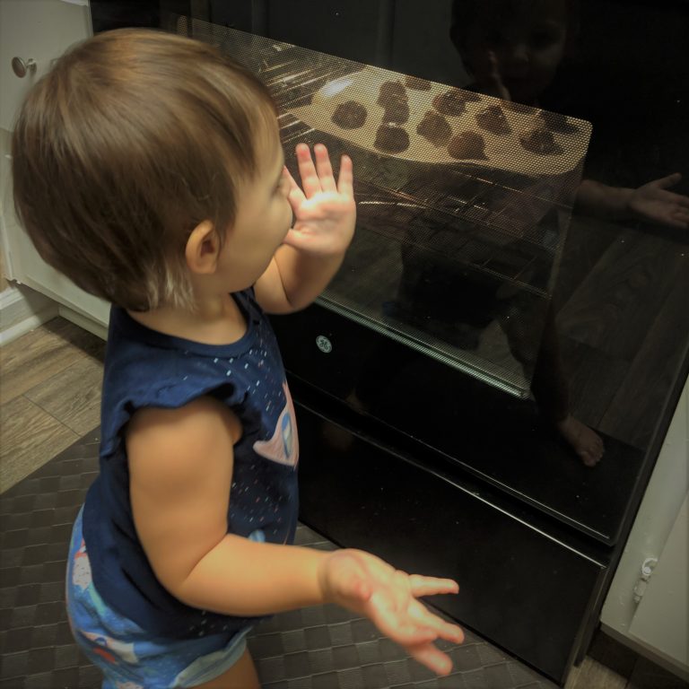 A baby looks into an oven with baking cookies in it.