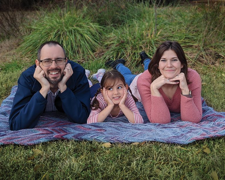A man, a girl, and a woman lay on a blanket in the grass with their hands under their chins.