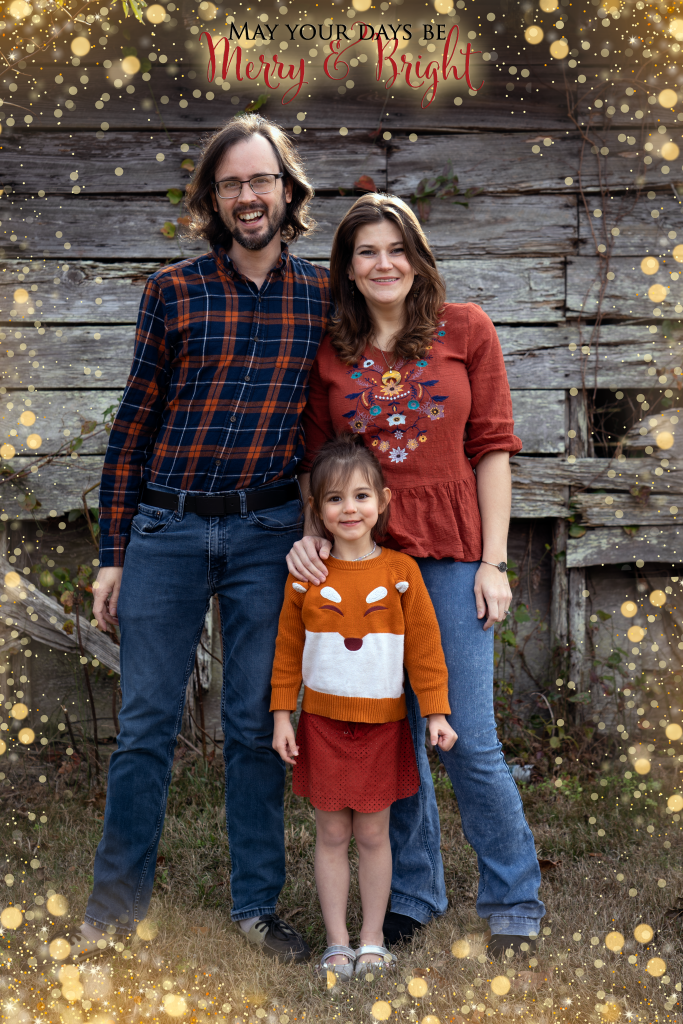 A man in blue and orange plaid. A woman in orange floral top. A girl in orange fox sweater and red skirt. Stand in front of an old wood wall.